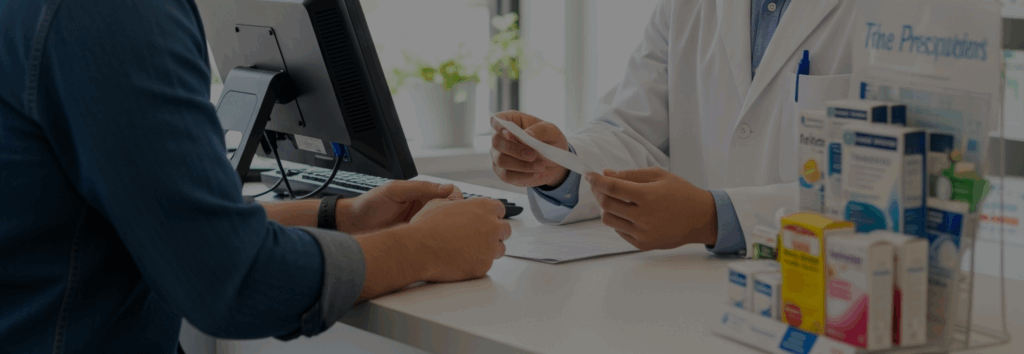 Um close-up de um farmacêutico em um jaleco branco segurando um papel enquanto conversa com um cliente, que está sentado em frente a um monitor de computador em um balcão de farmácia. Há caixas de medicamentos em exposição à direita.